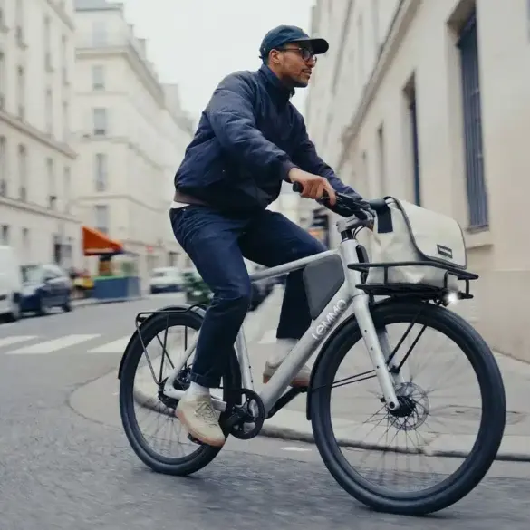 Ein Mann fährt ein silbernes Lemmo One "Collab. Ride" E-Bike mit einer Stofftasche über dem Vorderrad und einem Gepäckträger hinten durch eine Stadtstraße.