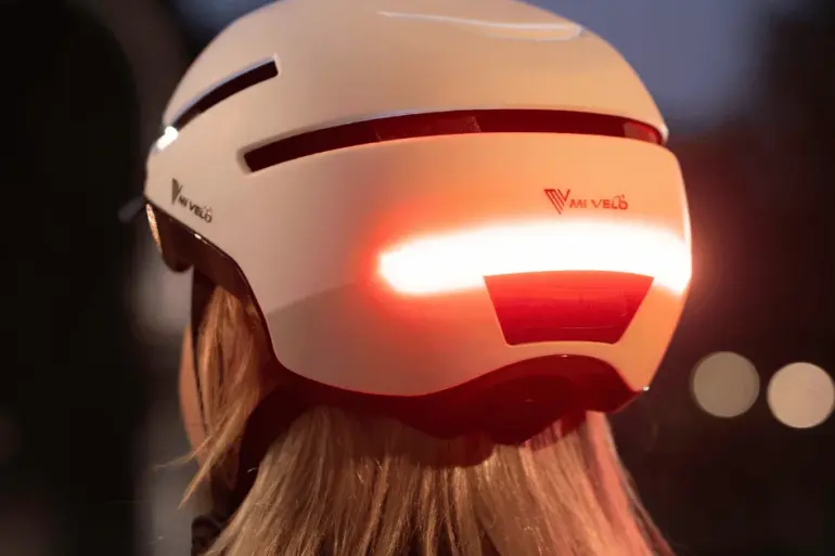 Fahrradhelm NOVA in Weiß mit aktiviertem rotem Rücklicht bei Dämmerung, gesehen von hinten auf dem Kopf einer Person mit blonden Haaren.