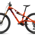 Orangefarbenes R.XX ULTIMATE Elektro-Mountainbike mit schwarzen Akzenten, goldener Federgabel und roten/weißen Logos auf dem Rahmen, präsentiert vor einem neutralen Hintergrund.
