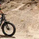 Mann fährt Rattan Pathfinder Pro E-Fatbike auf einem felsigen Bergpfad, Staub aufwirbelnd.