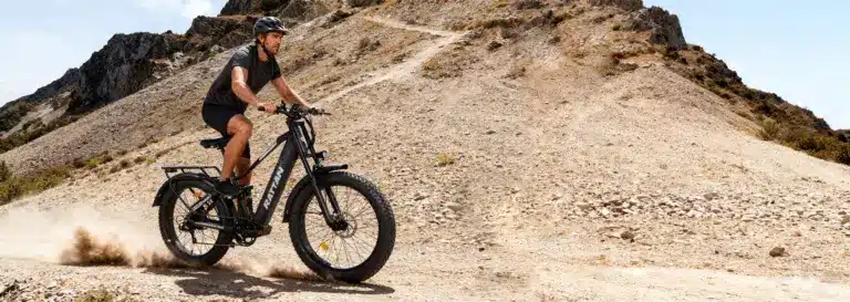 Mann fährt Rattan Pathfinder Pro E-Fatbike auf einem felsigen Bergpfad, Staub aufwirbelnd.