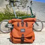 Orangefarbene ROCKBROS Lenkertasche für Brompton-Fahrräder, mit dunkelbraunen Schnallen und Henkeln, steht auf einem Steinfußboden vor einem grünen Brompton-Fahrrad.