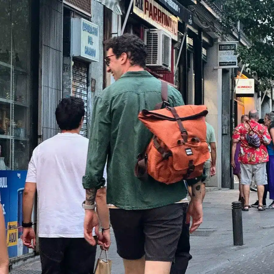 Mann mit orangefarbener Rucksacktasche, getragen auf einer Schulter, geht auf einer belebten Straße.