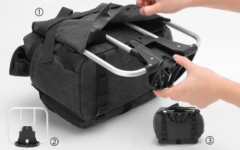 Nahaufnahme des schwarzen Befestigungsadapters, der an einer grauen Fahrradtasche mit Aluminiumrahmen montiert wird. Hände führen den Adapter ein.