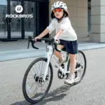 Mädchen in weißem Oberteil und dunkelblauer Hose fährt ein weißes ROCKBROS Kinder-Rennrad mit weißem Helm und Sportbrille.