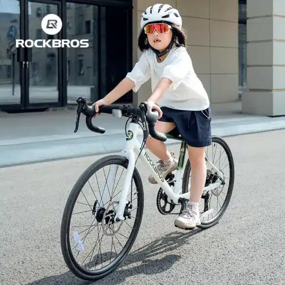 Mädchen in weißem Oberteil und dunkelblauer Hose fährt ein weißes ROCKBROS Kinder-Rennrad mit weißem Helm und Sportbrille.