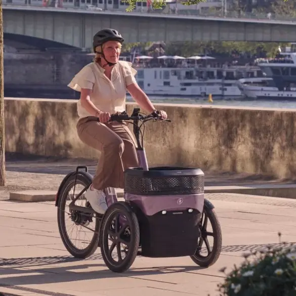 Eine Frau mit Helm fährt ein lila-graues Dreirad mit zwei Vorderrädern und einer großen Ladebox zwischen den Rädern.
