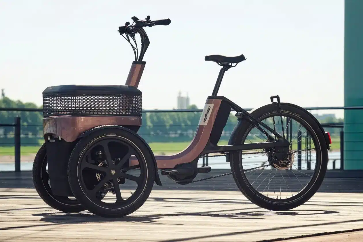 Ein rosegoldenes VanMoof Thuja Dreirad steht auf einem Holzdeck. Es hat zwei Räder vorne und ein Rad hinten. Vorne ist ein großer Korb angebracht.