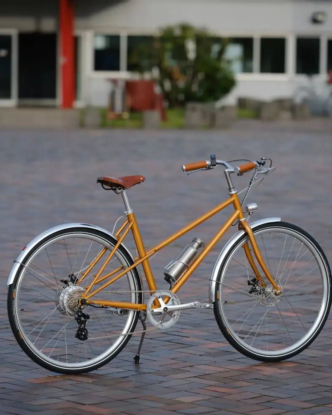 Goldenes Capri Azur Stinger Orange 4 E-Bike mit silbernen Schutzblechen und einem silbernen Flaschenhalter, stehend auf einem gepflasterten Hintergrund.