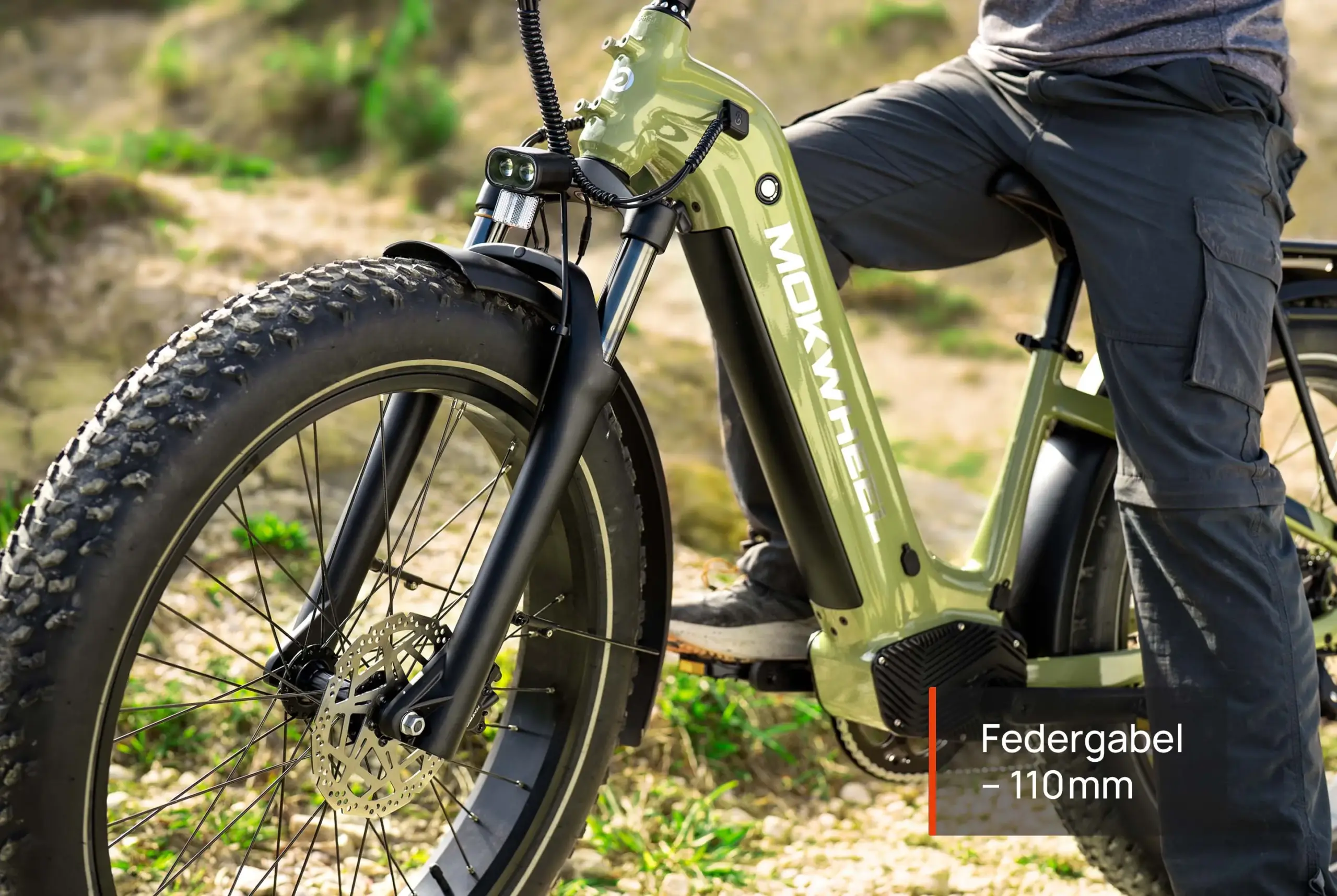 Detailansicht des grünen Basalt ST 2.0 E-All-Terrain-Bikes mit breiten Reifen, hydraulischen Scheibenbremsen und einer Federgabel von 110 mm, während eine Person darauf sitzt.