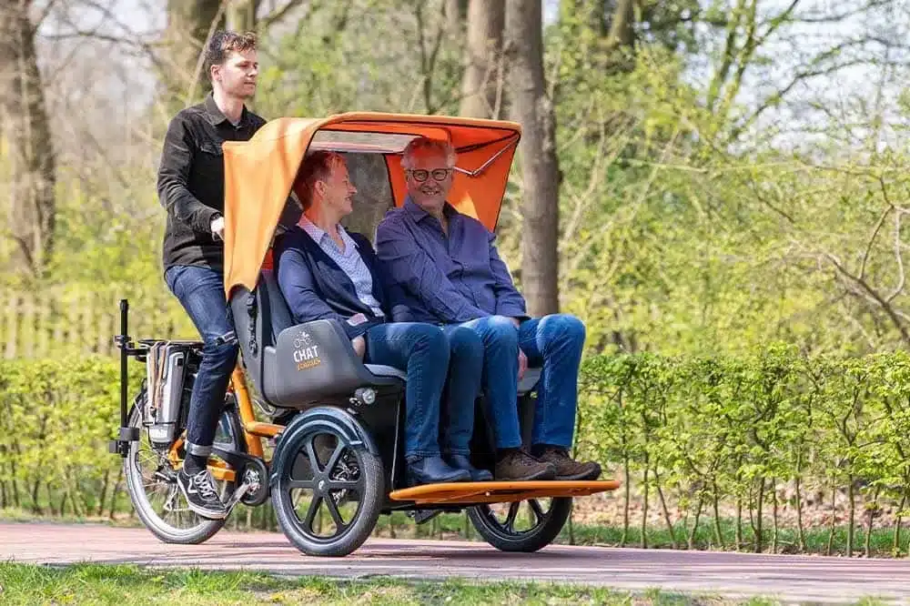 Dreirädriges Van Raam Chat Fahrrad mit Sitzplatz für zwei Personen, das von einer Person gefahren wird und auf einem Weg durch eine grüne Parklandschaft unterwegs ist.