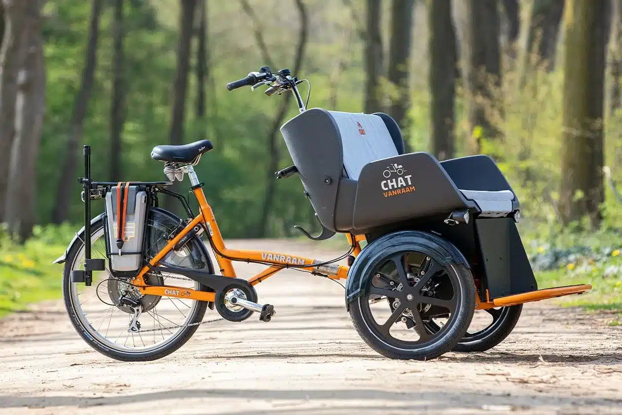 Ein oranges dreirädriges Van Raam Chat Fahrrad mit integriertem Sitz für einen Passagier, fotografiert im Freien auf einem Feldweg.