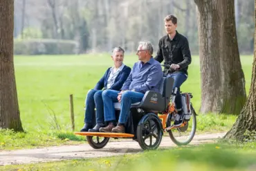 Zwei ältere Menschen, eine Frau und ein Mann, sitzen nebeneinander auf einem speziellen Fahrrad, das für den Transport von Personen konzipiert ist. Ein Mann fährt das Fahrrad von hinten. Das Fahrrad hat ein dunkles Sitzmodul mit dem Aufdruck "CHAT VAN RAAM". Die Fahrt findet auf einem Feldweg statt, umgeben von Grünflächen und Bäumen.