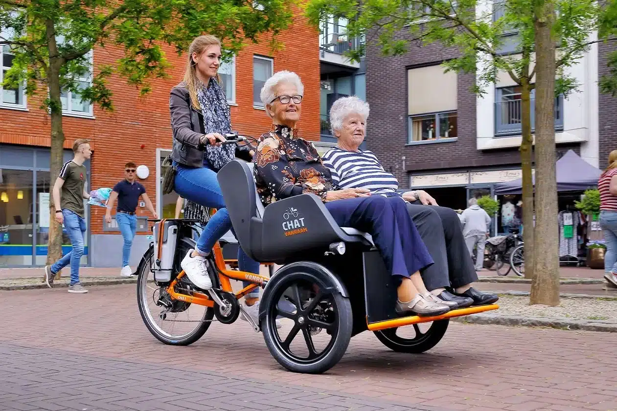 Eine junge Frau fährt auf einem orangen Van Raam Chat Dreirad. Auf der Rückbank sitzen zwei ältere Damen nebeneinander. Das Fahrrad ist auf einer gepflasterten Straße in einer Stadt unterwegs.