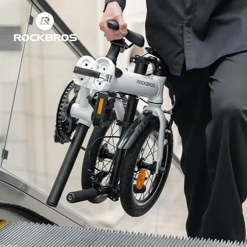 Ein Mann trägt das zusammengeklappte weiße ROCKBROS IFN 14-Zoll-Faltrad auf einer Rolltreppe. Das Fahrrad ist kompakt und für den urbanen Gebrauch konzipiert.
