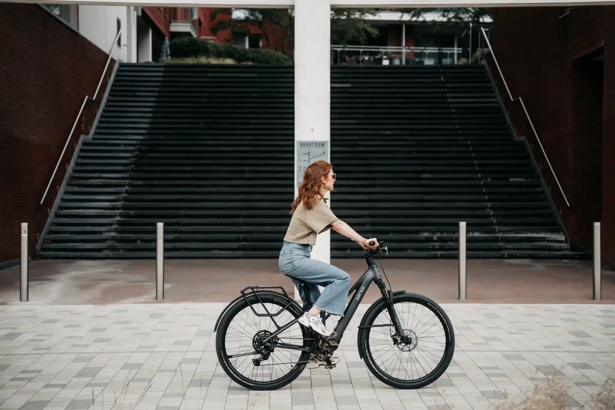 Frau mit roten Haaren fährt ein schwarzes Aeres Leuven SX Mid Step E-Bike auf einem gepflasterten Weg vor einer großen Treppe.
