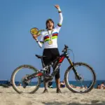 Die Radsportlerin Helen Weber steht mit ihrem ROTWILD R.EXC E-Enduro-Bike am Strand. Sie trägt das Weltmeistertrikot und hält ihre Medaille und eine Trophäe in den Händen. Ihr Arm ist siegessicher erhoben.