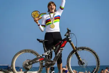 Die Radsportlerin Helen Weber steht mit ihrem ROTWILD R.EXC E-Enduro-Bike am Strand. Sie trägt das Weltmeistertrikot und hält ihre Medaille und eine Trophäe in den Händen. Ihr Arm ist siegessicher erhoben.