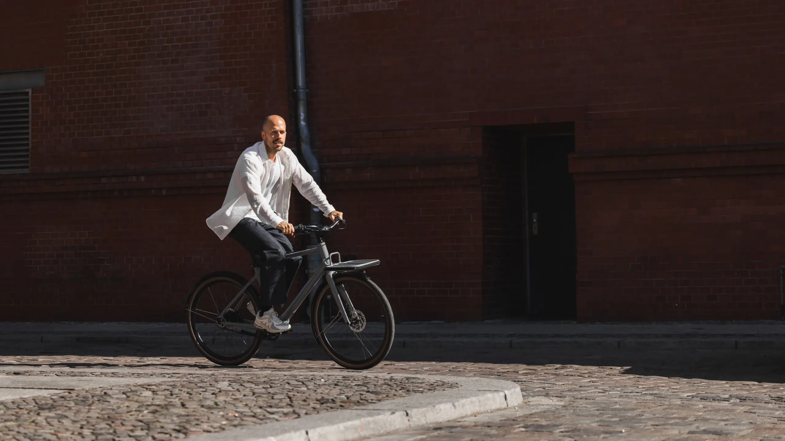 Ein Mann mit kurzgeschorenen Haaren und Bart fährt ein modernes, graues Schindelhauer Emil E-Bike auf einem historischen Kopfsteinpflasterweg. Er trägt ein weißes, leichtes Hemd über einem dunklen T-Shirt und dunkle Hosen. Das Fahrrad ist mit einem vorderen Gepäckträger ausgestattet. Im Hintergrund ist eine dunkelrote Backsteinwand zu sehen.
