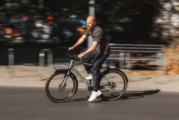Ein Mann fährt mit einem grauen Schindelhauer Emil E-Bike mit Gepäckträger auf einer asphaltierten Straße, die Hintergrundelemente sind durch die Bewegung verschwommen.