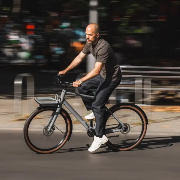 Ein Mann fährt mit einem grauen Schindelhauer Emil E-Bike mit Gepäckträger auf einer asphaltierten Straße, die Hintergrundelemente sind durch die Bewegung verschwommen.