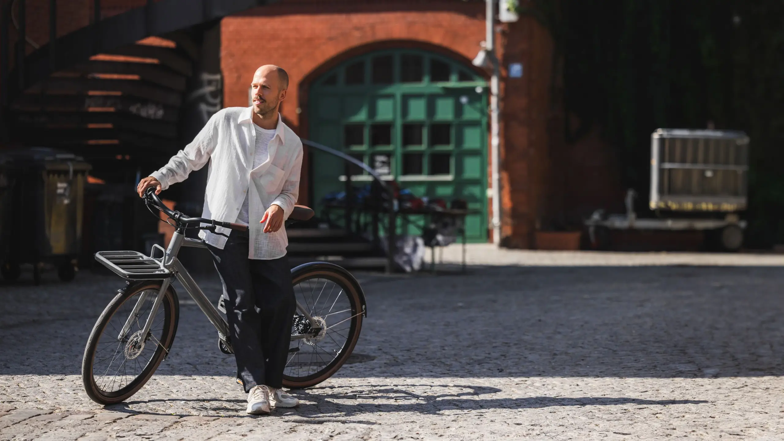 Ein Mann mit weißem Hemd und dunkler Hose lehnt lässig an einem grauen Schindelhauer Emil E-Bike. Im Hintergrund ist ein Backsteinhaus mit einem grünen Tor zu sehen.