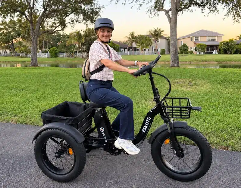 Eskute T302 Pro Elektro-Lastendreirad mit breiten Reifen und Körben vorne und hinten, gefahren von einer Frau mit Helm.