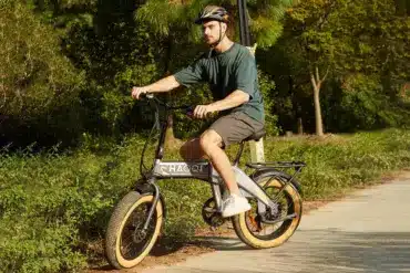 Mann mit Helm fährt ein silbernes Haoqi Squirrel faltbares E-Bike mit breiten Fat-Reifen auf einem asphaltierten Weg, umgeben von grüner Vegetation.