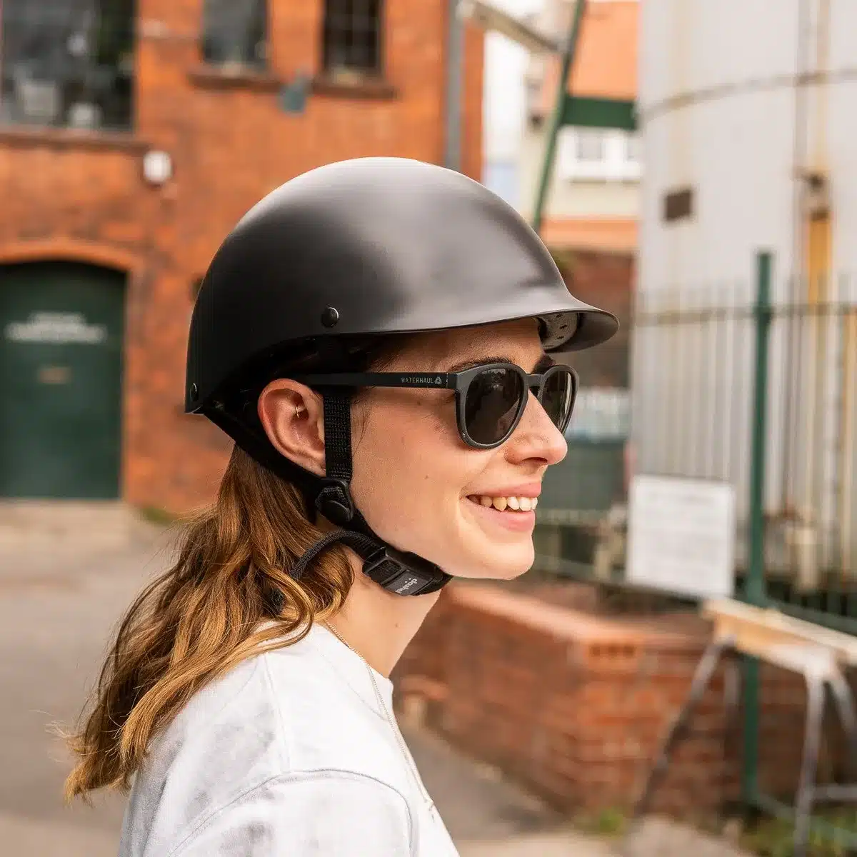 Frau mit seitlichem Blick trägt einen mattschwarzen City-Helm und eine Sonnenbrille, unterstrichen durch die urbane Umgebung.