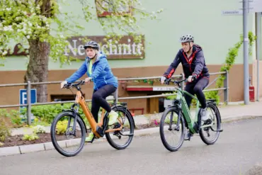 Zwei Personen mit Helm fahren auf E-Trekkingbikes durch eine Straße, die von Bäumen gesäumt ist.