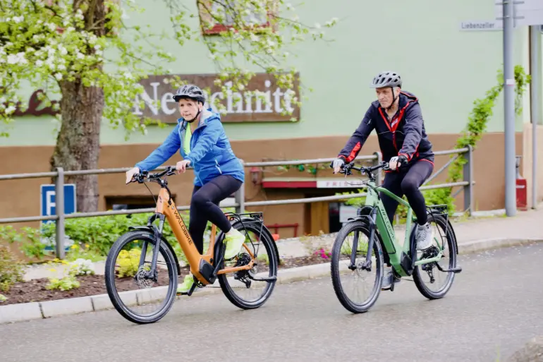 Zwei Personen mit Helm fahren auf E-Trekkingbikes durch eine Straße, die von Bäumen gesäumt ist.