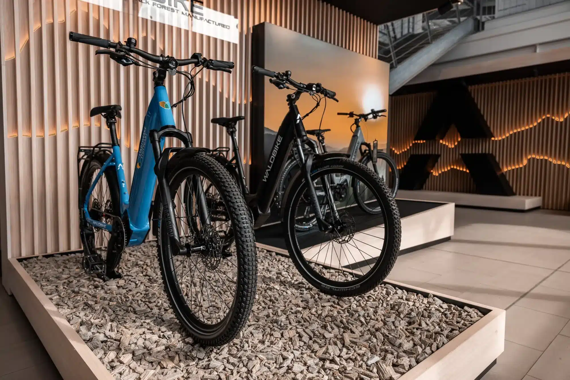 Zwei E-Trekkingbikes, ein blaues und ein schwarzes, werden auf einem Kiesbett stehend ausgestellt.