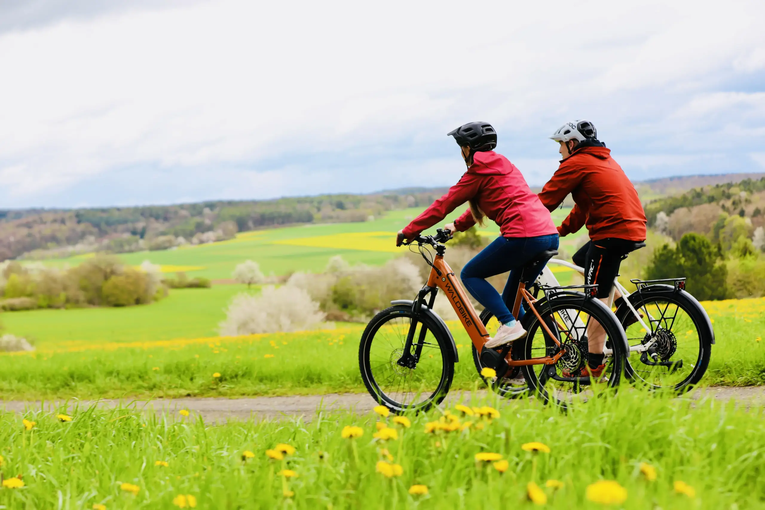 Zwei Radfahrer mit Helmen fahren auf E-Bikes auf einem Feldweg in idyllischer Natur. Der Hintergrund zeigt grüne Hügel und blühende Bäume.